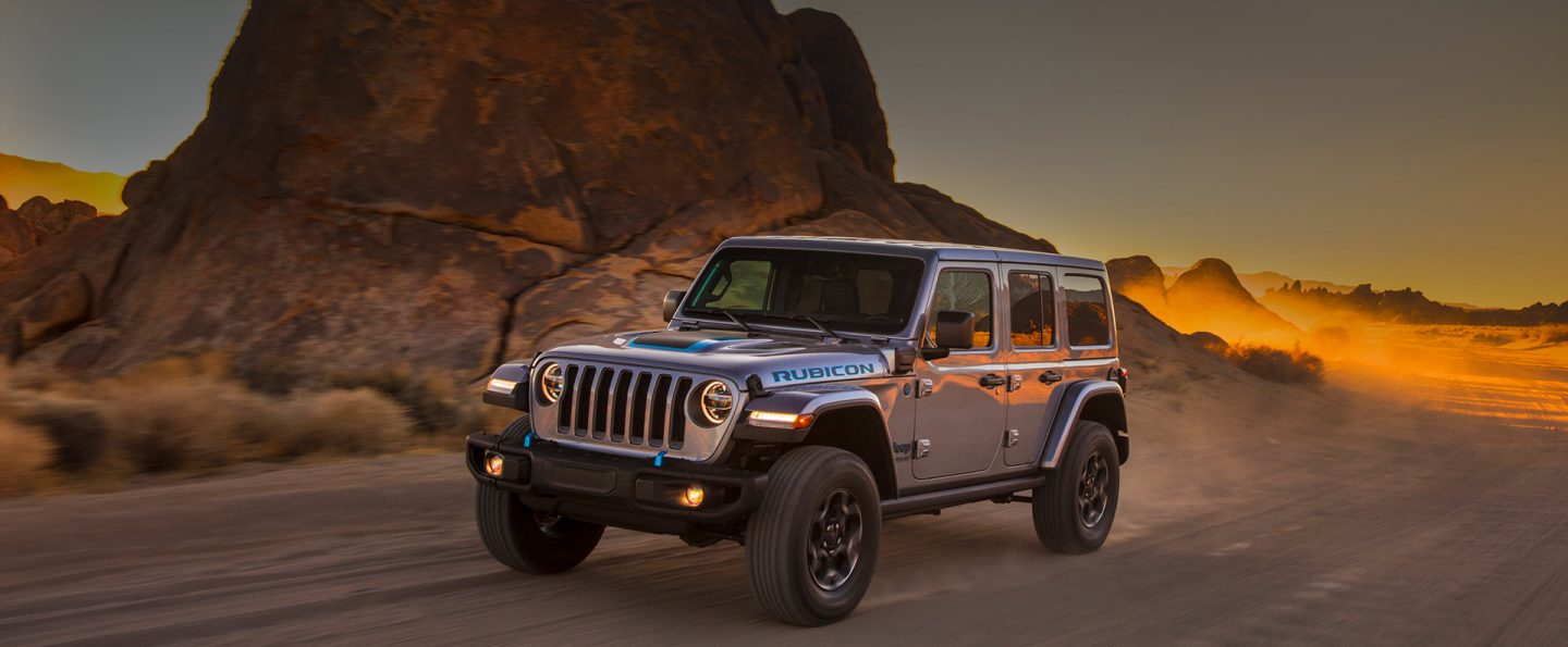 Three-quarter side view of a 2021 Jeep Wrangler 4xe being driven through a dusty mountainous area at sunset.