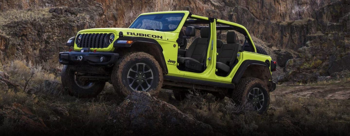 Dos modelos de Jeep Wrangler estacionados en un claro rocoso en las montañas. A la izquierda, un Wrangler 4xe Rubicon gris de cuatro puertas. A la derecha, un Wrangler Willys gris de dos puertas.