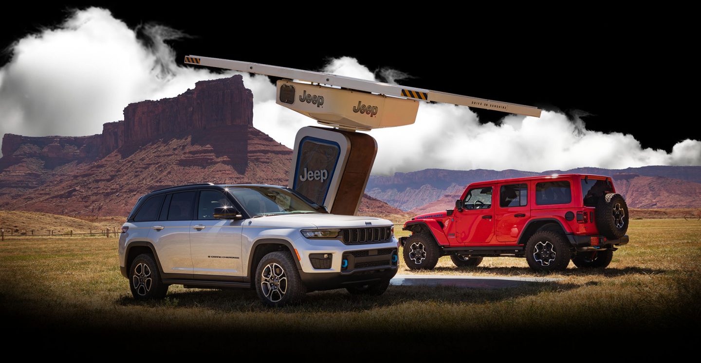 Un Jeep Wrangler Rubicon 4xe y un Jeep Grand Cherokee Trailhawk 4xe 2022 estacionados en una estación de carga rodeada de montañas.